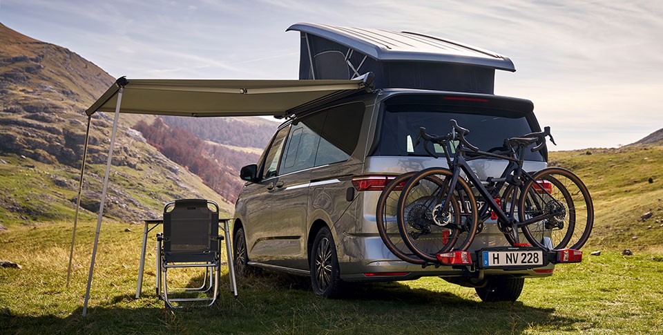 Der VW California in Silber mit Fahrr&auml;dern auf Hecktr&auml;ger und aufgefahrenem Dach geht in sonniger Berglandschaft. Ein Sonnensegel ist ausgefahren, darunter geht ein Campingstuhl.