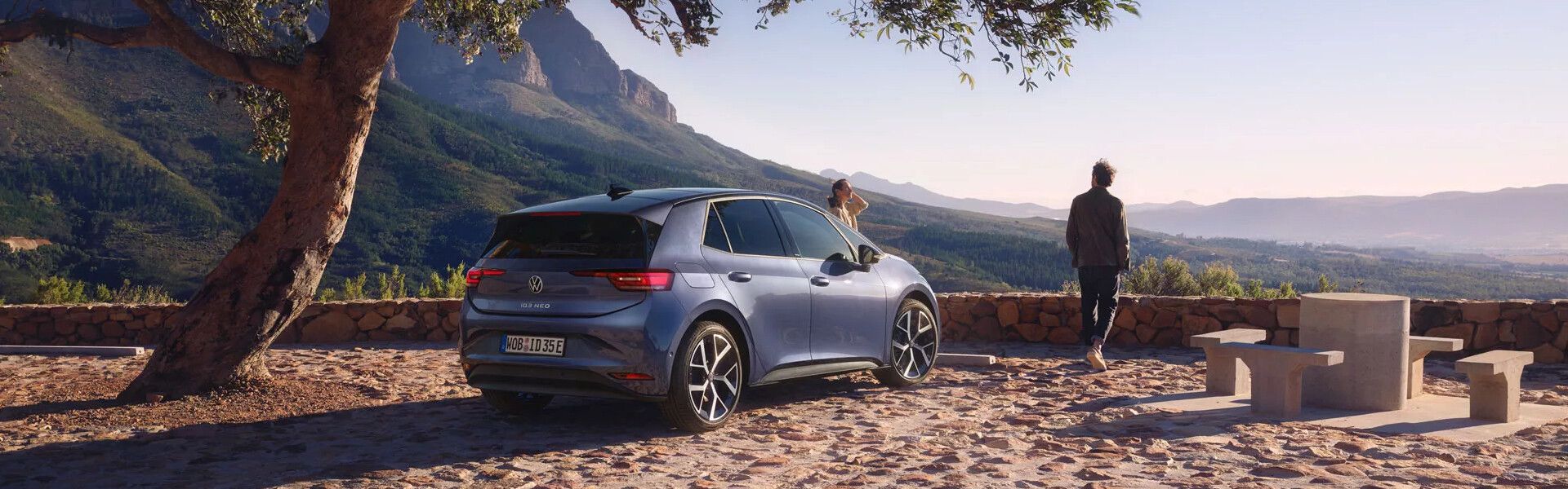 Heckansicht des neuen ID.3 Neo in Dunkelblau auf mediterranem Aussichtspunkt mit Blick in die waldige Weite. Zwei Personen stehen vor dem Fahrzeug.