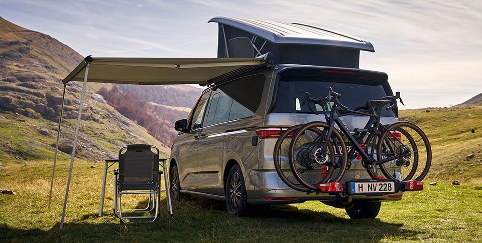 Der VW California in Silber mit Fahrr&auml;dern auf Hecktr&auml;ger und aufgefahrenem Dach geht in sonniger Berglandschaft. Ein Sonnensegel ist ausgefahren, darunter geht ein Campingstuhl.
