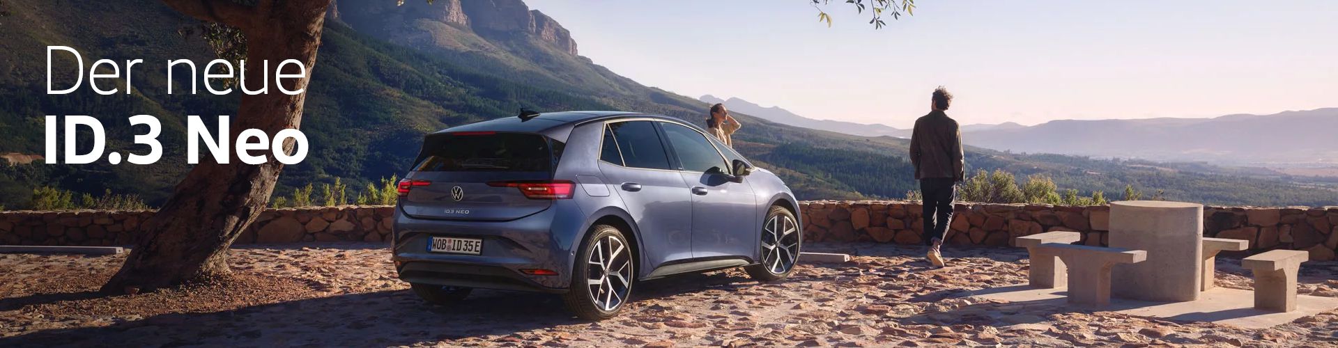 Heckansicht des neuen ID.3 Neo in Dunkelblau auf mediterranem Aussichtspunkt mit Blick in die waldige Weite. Zwei Personen stehen vor dem Fahrzeug. Text: Der neue ID.3 Neo