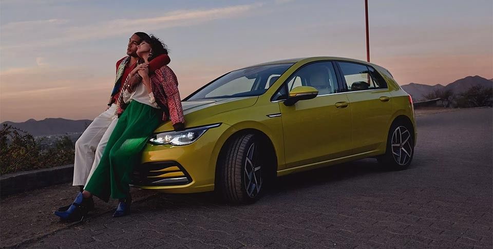 Mann und Frau lehnen auf Motorhaube von gelbem VW Golf und schauen in Richtung Sonnenuntergang