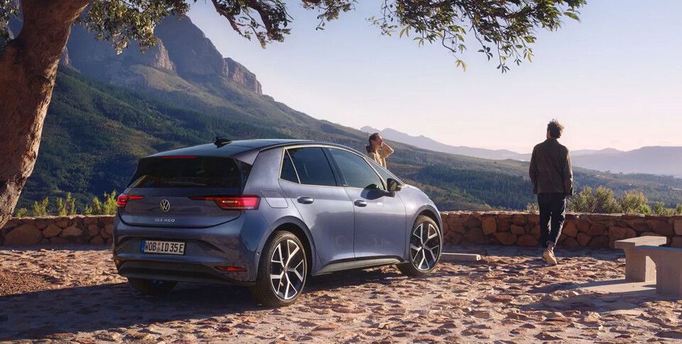 Heckansicht des neuen ID.3 Neo in Dunkelblau auf mediterranem Aussichtspunkt mit Blick in die waldige Weite. Zwei Personen stehen vor dem Fahrzeug.