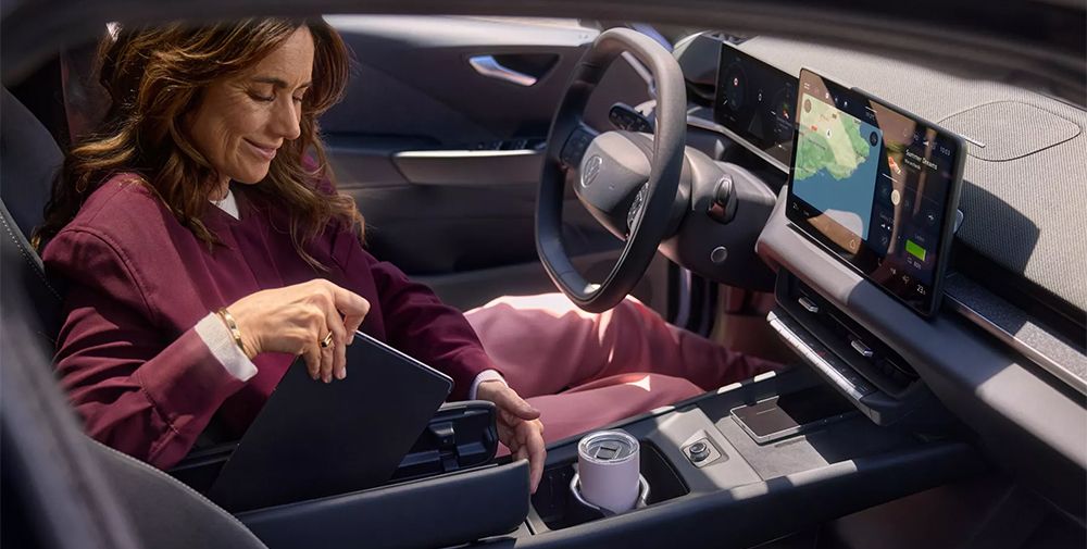 Interieur/Cockpit des ID.3 Neo. Frau in r&ouml;tlichem Anzug sitzt auf Fahrersitz und verstaut ein Tablet in der Mittelarmst&uuml;tze.